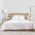 Minimalistická spálňa s bielymi stenami, drevenou posteľou, dvoma bielymi nočnými stolíkmi a bielou otvorenou policovou zostavou.