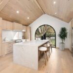 Svetlá kuchyňa so šikmým dreveným stropom, drevenými skrinkami a bielym ostrovčekom pred oblúkovým oknom.