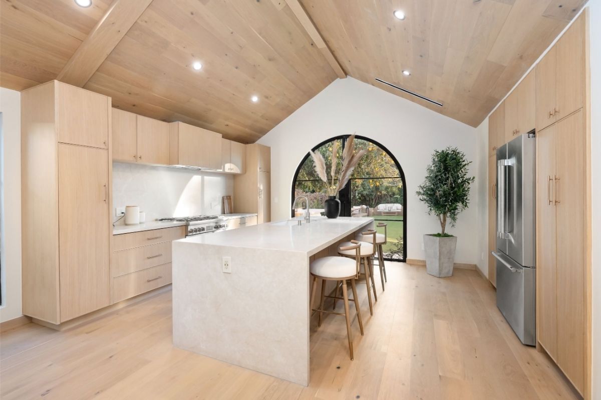Svetlá kuchyňa so šikmým dreveným stropom, drevenými skrinkami a bielym ostrovčekom pred oblúkovým oknom.