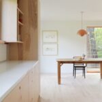 Interiér spájajúci kuchyňu a jedáleň so svetlými drevenými podlahami, jednoduchým dreveným stolom a oranžovou lampou.