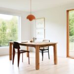 ednoduchý jedálenský priestor s dreveným stolom, dvomi stoličkami a veľkými oknami s výhľadom do záhrady.