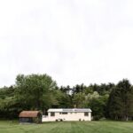 Nenápadný svetlý rodinný dom s hnedou hospodárskou budovou na širokej zelenej lúke pri lese.