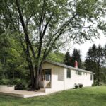 Prízemný drevený dom s terasou a vonkajším schodiskom pod košatým stromom uprostred záhrady.