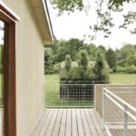 Drevená terasa s bielym mriežkovým zábradlím s výhľadom na hospodársku budovu a zeleň.