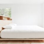 Spálňa s bielou stenou, drevenou podlahou a jednoduchou platformovou posteľou so vstavaným čelom.