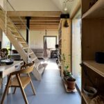 Svetlý moderný interiér s priznanými drevenými trámami, schodiskom, šedou podlahou a regálmi s rastlinami.