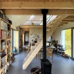 Otvorený obytný priestor s knižnicami, dreveným schodiskom, čiernym krbom a veľkými oknami s výhľadom na zeleň.