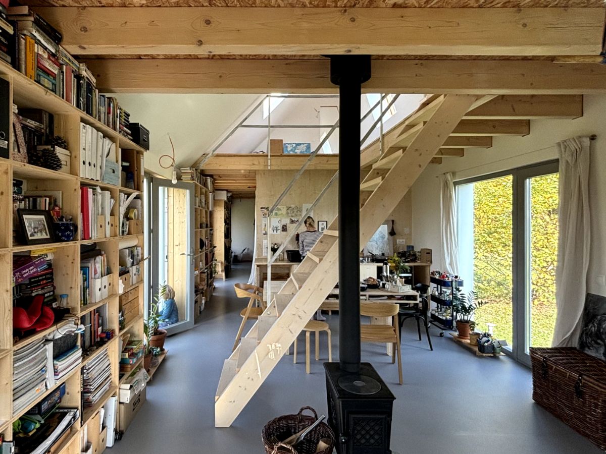 Otvorený obytný priestor s knižnicami, dreveným schodiskom, čiernym krbom a veľkými oknami s výhľadom na zeleň.