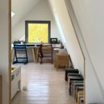 Podkrovná izba s bielymi šikmými stenami, svetlou drevenou podlahou a pracovným stolom pri okne s výhľadom na žltú farbu jesene.