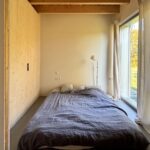 Spálňa s dreveným , trámovým stropom, posteľou s tmavosivou prikrývkou a vysokým oknom so závesmi.