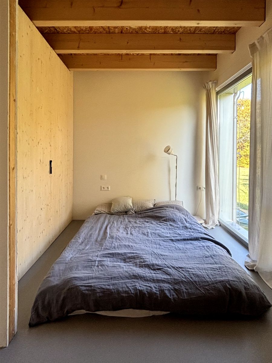 Spálňa s dreveným , trámovým stropom, posteľou s tmavosivou prikrývkou a vysokým oknom so závesmi.