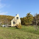 Predná strana domu s dreveným obkladom, šikmou strechou a dvoma čiernymi oknami uprostred trávnatej plochy,