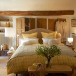 Vidiecka spálňa so žlto-bielou károvanou posteľou, dreveným trámom a terakotovou dlažbou.