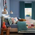 Vidiecka spálňa so žlto-bielou károvanou posteľou, dreveným trámom a terakotovou dlažbou.