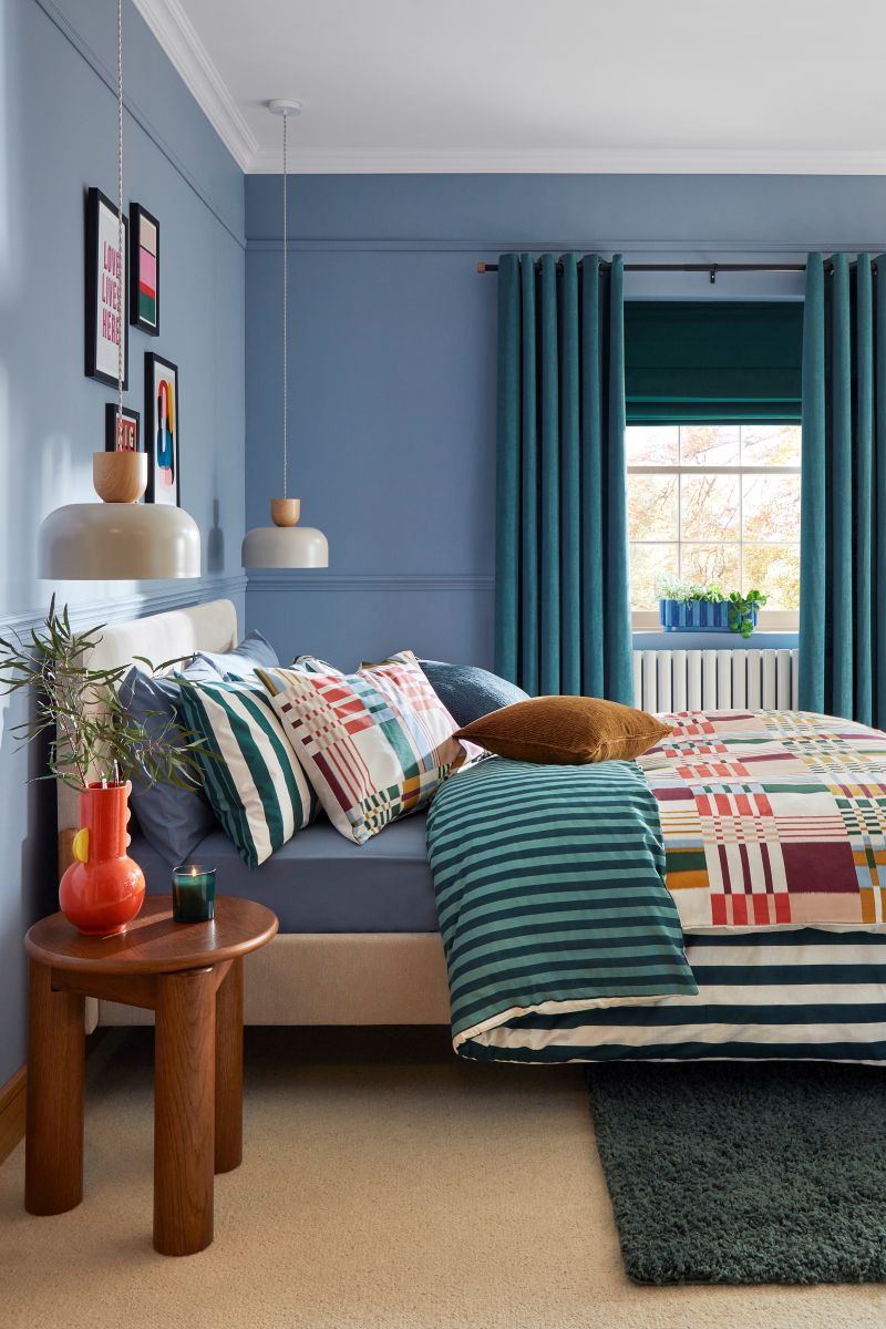 Vidiecka spálňa so žlto-bielou károvanou posteľou, dreveným trámom a terakotovou dlažbou.