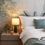 Spálňa s bielou posteľou, sivým nočným stolíkom a tapetou s motívom zelených palmových listov.