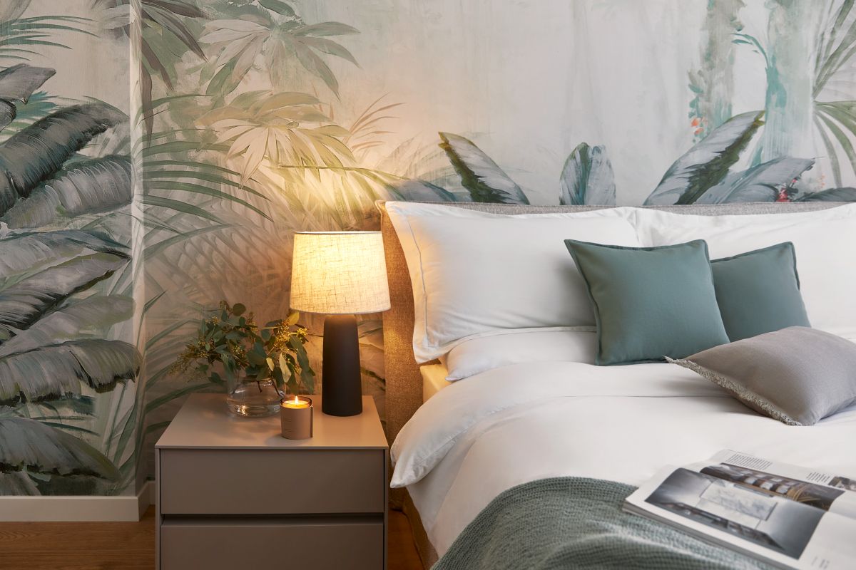 Spálňa s bielou posteľou, sivým nočným stolíkom a tapetou s motívom zelených palmových listov.