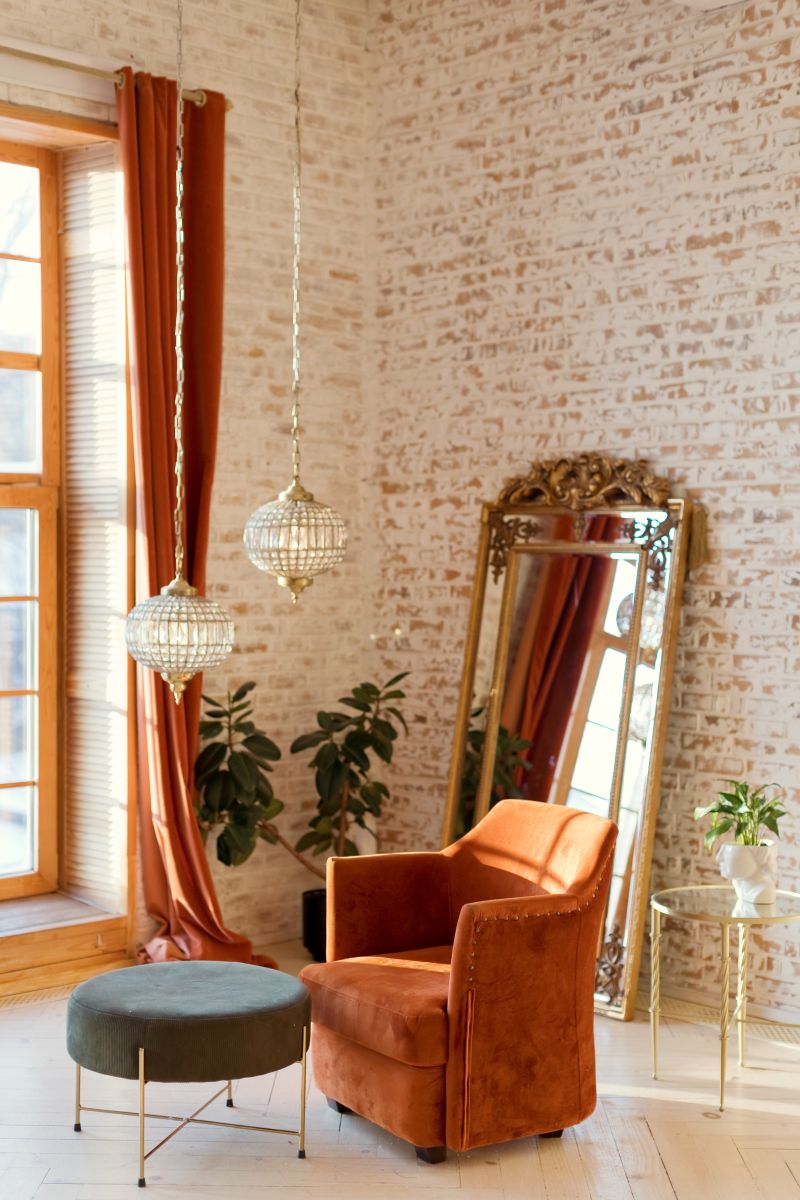 Terakotové zamatové kreslo pri tehlovej stene vedľa rastlín a opretého zrkadla.