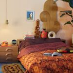 Posteľ v spálni v sýtych odtieňoch fialovej oranžovej a terakotovej, drevený nočný stolík.