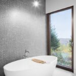 Kúpeľňa s bielou voľne stojacou vaňou, sivou mozaikou na stene a vysokým oknom s výhľadom do prírody.