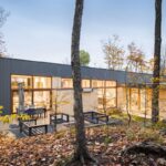 Moderný nízky dom s drevenou terasou s vonkajším sedením a s tmavým a dreveným obkladom.