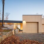 Príjazdová cesta a moderná fasáda domu s drevenými garážovými dverami a kombináciou dreveného a tmavosivého obkladu.