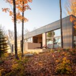 Moderný dom s terasou s drevenými lamelami, svetlým dreveným a tmavosivým obkladom uprostred jesenných stromov.