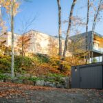 Exteriér domu na svahu s tmavosivou garážou v popredí a drevenou hlavnou stavbou osadenou v skalnatom teréne.