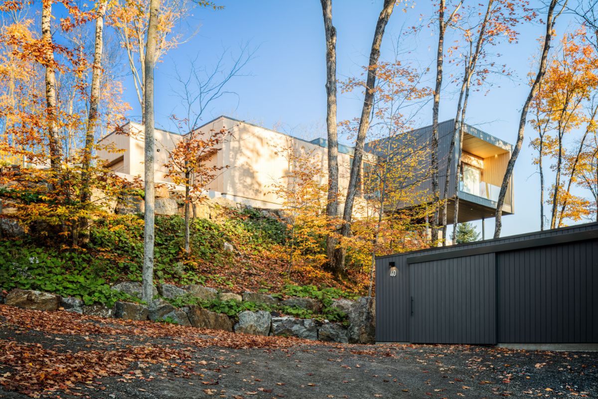 Exteriér domu na svahu s tmavosivou garážou v popredí a drevenou hlavnou stavbou osadenou v skalnatom teréne.