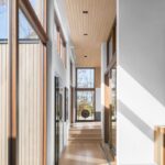 Dlhá minimalistická chodba s vysokými drevenými oknami, svetlou drevenou podlahou a lamelovým dreveným stropom.