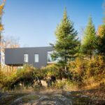 Moderná, hranatá stavba domu s tmavosivou fasádou posadená na skalnatom teréne a obklopená jesennou vegetáciou.