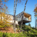 Moderný hranatý dom s vysunutým tmavosivým kubusom a drevenou fasádou na svahu uprostred jesenného lesa.