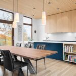 Moderná kuchyňa a jedáleň s veľkým dreveným stolom, tmavomodrým ostrovom a hornými skrinkami vo farbe prírodného dreva.