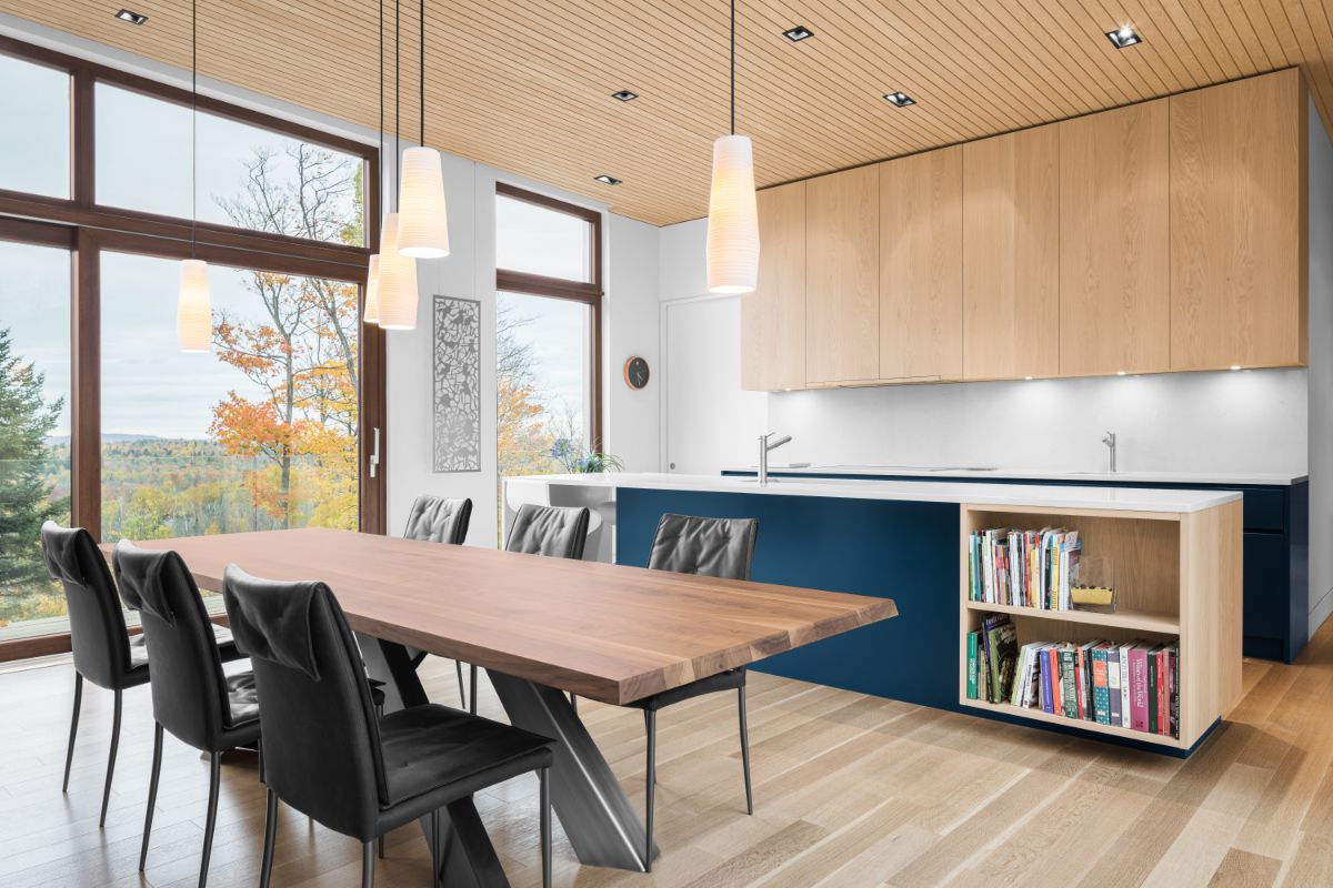 Moderná kuchyňa a jedáleň s veľkým dreveným stolom, tmavomodrým ostrovom a hornými skrinkami vo farbe prírodného dreva.