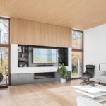 Moderná obývacia izba s dreveným obkladom steny, integrovaným TV a krbom a veľkými oknami do jesennej prírody.