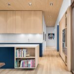 Minimalistická kuchyňa s modrým ostrovom, svetlými drevenými hornými skrinkami a lamelovým dreveným stropom.