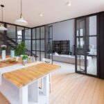 Pohľad z jedálne s jedálenským stolom do obývačky, oddelenej sklenenou priečkou so skladacími dverami.