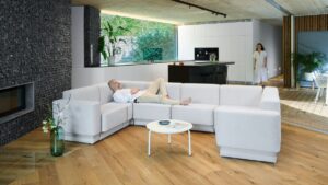 Muž leží na viacmiestnej sedačke do U pri kuchyni v modernom interiéri, žena k nemu kráča cez kuchyňu.