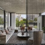 Luxusná moderná obývacia izba s béžovou zamatovou sedačkou, betónovým stropom a vysokými presklenými oknami do záhrady.