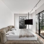 Svetlá spálňa s bielou stenou, veľkou čalúnenou posteľou a presklenou stenou s lamelovým dekorom.