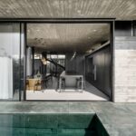 Pohľad z exteriéru na bazén a otvorenú modernú jedáleň a kuchyňu s betónovým stropom a tmavými prvkami.