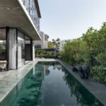 Dlhé úzke vonkajšie jazierko s tmavým obkladom pri vilke so živým plotom a modernou sochou psa.