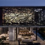 Panoramatický nočný pohľad na vilu s veľkým otvoreným preskleným priestorom a dekoratívnym lamelovým obkladom.