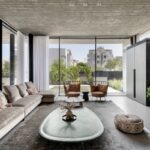 Priestranná obývacia izba s béžovou zamatovou sedačkou, industriálnym betónovým stropom a veľkými oknami s výhľadom na okolie.