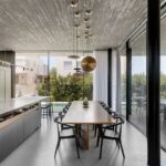 Moderná jedáleň s dreveným stolom a čiernymi stoličkami pod betónovým stropom, osvetlená dizajnovými lampami a denným svetlom.