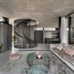 Priestranná obývacia izba s industriálnym betónovým stropom, svetlou zamatovou sedačkou a čiernym točitým schodiskom z kovu.