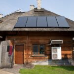 Pohľad na tradičnú drevenú chatu s trstinovou strechou s tmavými solárnymi kolektormi a vonkajšou jednotkou tepelného čerpadla.