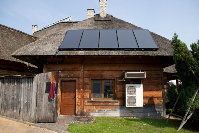 Pohľad na tradičnú drevenú chatu s trstinovou strechou s tmavými solárnymi kolektormi a vonkajšou jednotkou tepelného čerpadla.