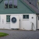 Moderné biele tepelné čerpadlo s okrúhlym ventilátorom inštalované na bielej fasáde domu so zeleným dreveným obkladom.