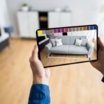 Ruky držiace tablet s aplikáciou rozšírenej reality na vizualizáciu sivej pohovky a farieb v izbe.